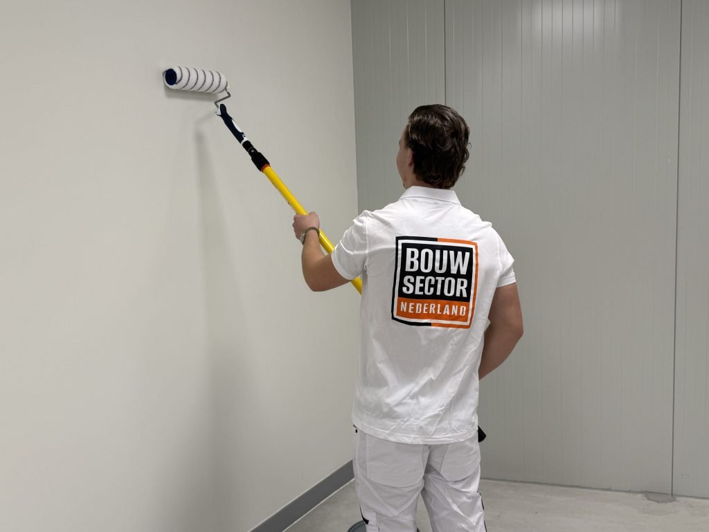 Vakman in witte werkkleding rolt een gladde witte wand met een roller aan een lange steel