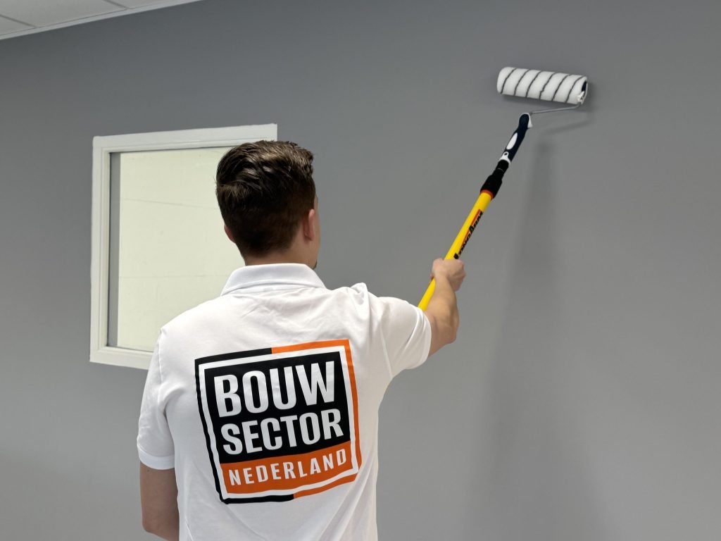 Schilder van Bouwsector Nederland rolt grijze verf op de muur naast een binnenraam met wit kozijn