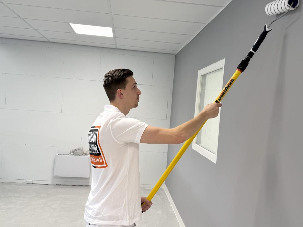 Schilder zet met een lange roller grijze muurverf bovenaan de wand bij het raam