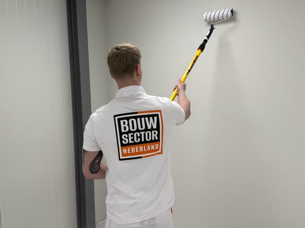 Schilder met logo van Bouwsector Nederland rolt verf op een lichte wand in een smalle ruimte