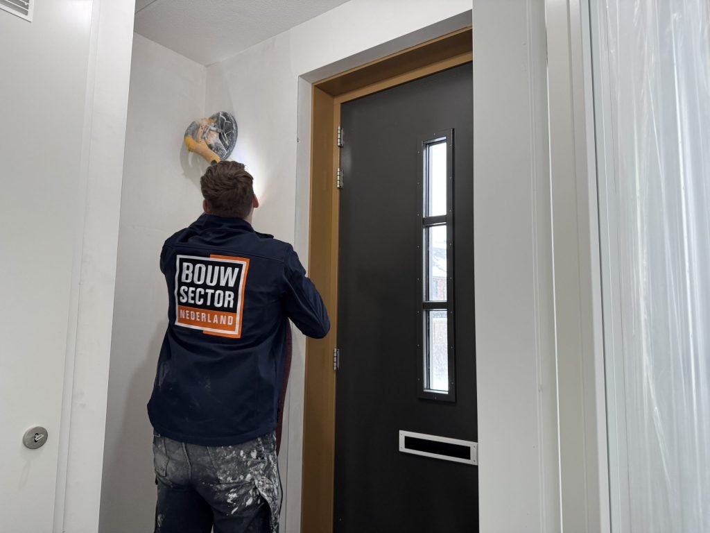 Vakman van Bouwsector Nederland schuurt met een wandschuurmachine de aansluiting van wand en plafond in een hal naast de voordeur